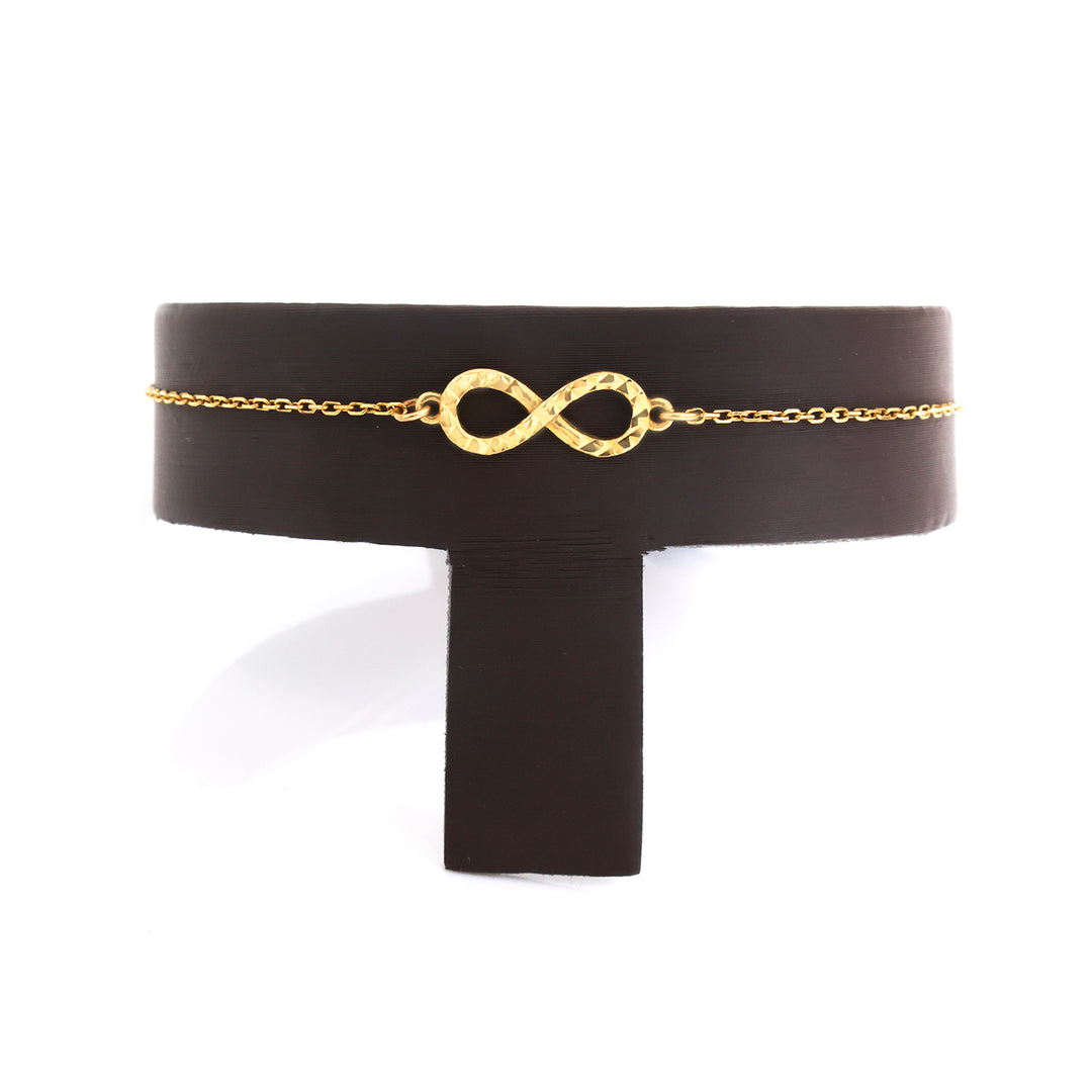 A delicate gold chain bracelet with a textured infinity symbol centerpiece, displayed on a black T-stand against a white background.