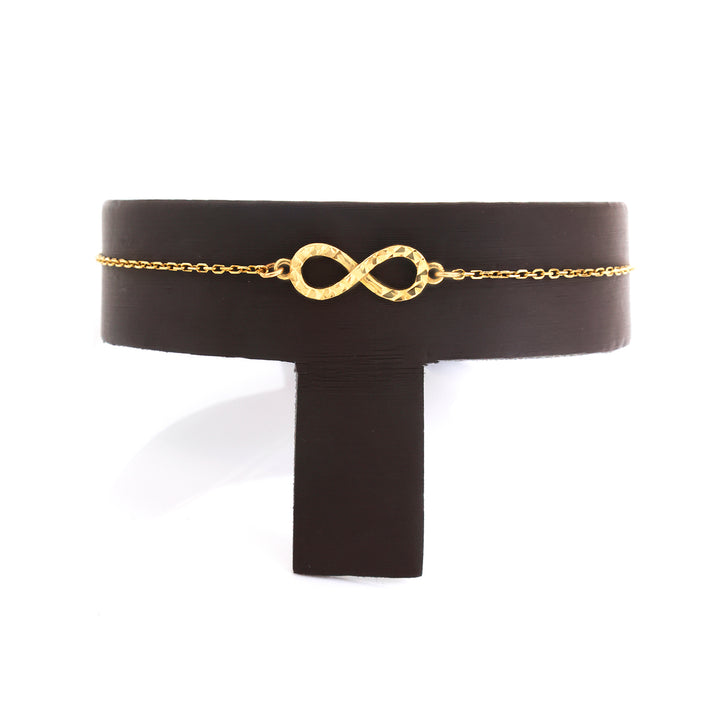 A delicate gold chain bracelet with a textured infinity symbol centerpiece, displayed on a black T-stand against a white background.