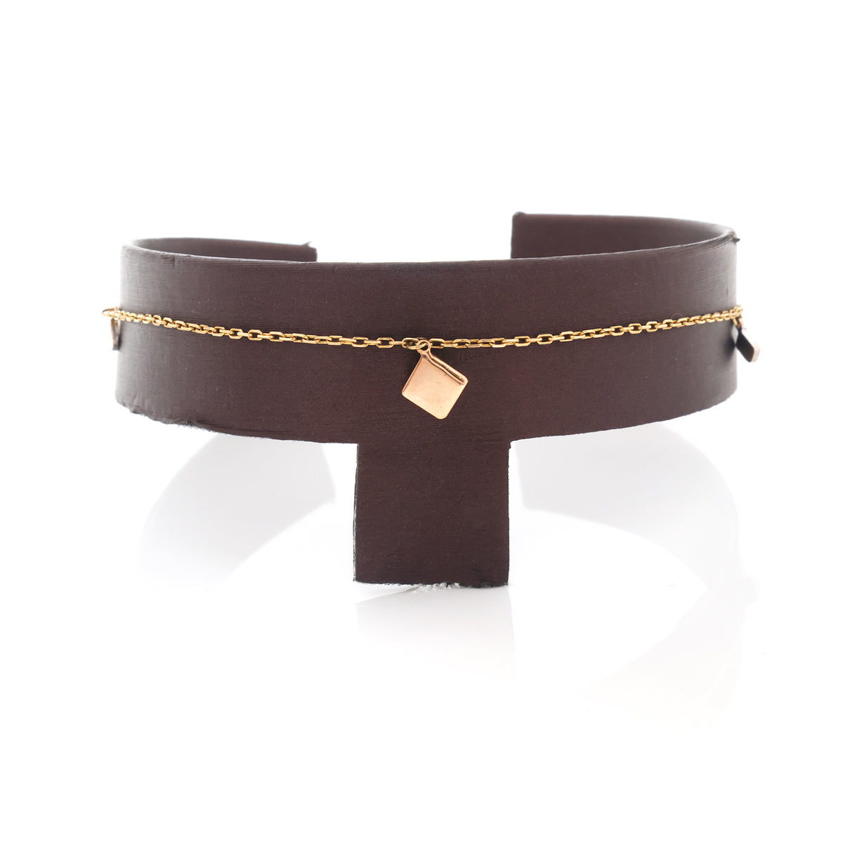A dark brown leather cuff bracelet with a delicate gold chain and a small rose gold cube charm, displayed on a white reflective surface.