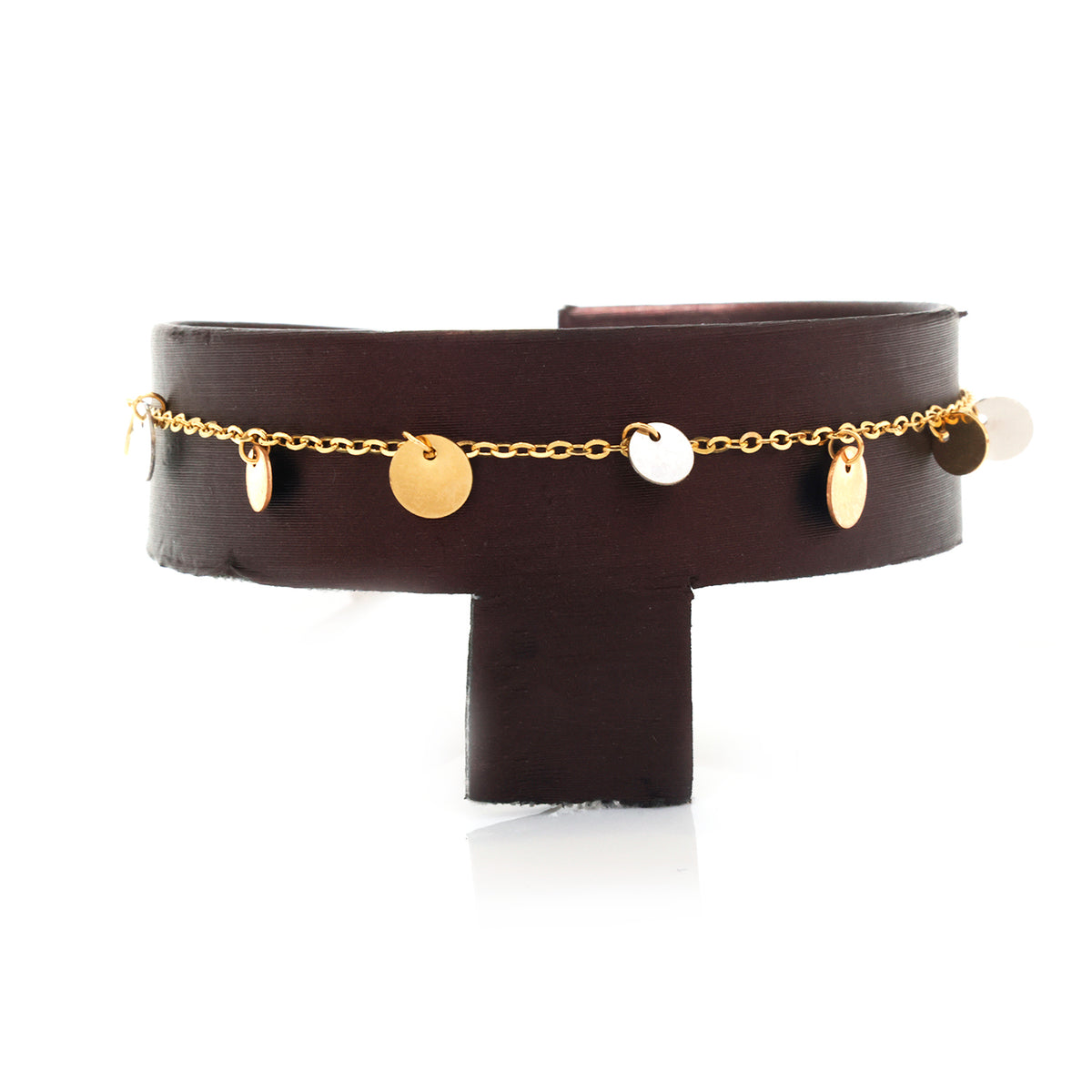 A dark brown leather cuff bracelet with a delicate gold chain draped across the front, adorned with small gold and silver disc charms, displayed on a black stand against a white background.