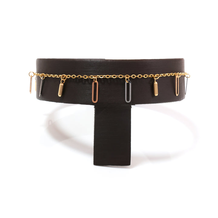 A wide black belt with a thin gold chain running around the waist and several dangling bar-shaped charms in gold, rose gold, and silver, displayed on a black stand against a white background.