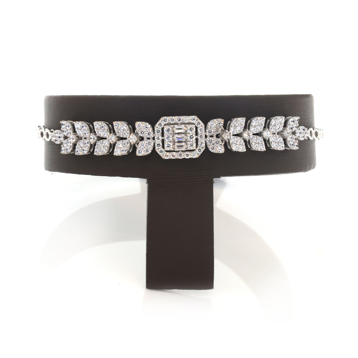A wide black cuff bracelet with a sparkling silver-toned geometric and leaf pattern, displayed on a black stand against a white background.