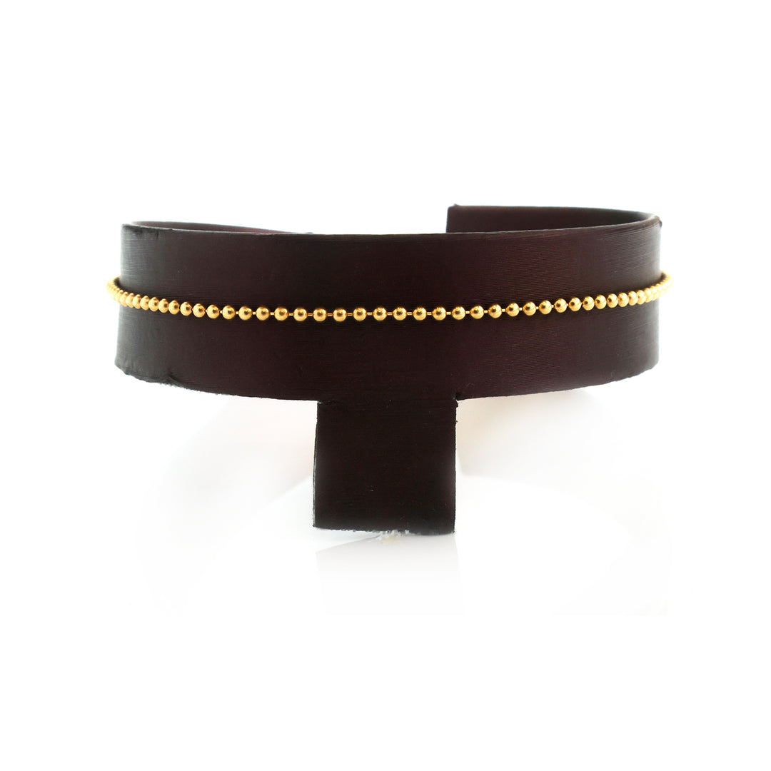 A wide black leather cuff bracelet featuring a delicate gold beaded chain running horizontally across the front, displayed on a black stand against a white background.