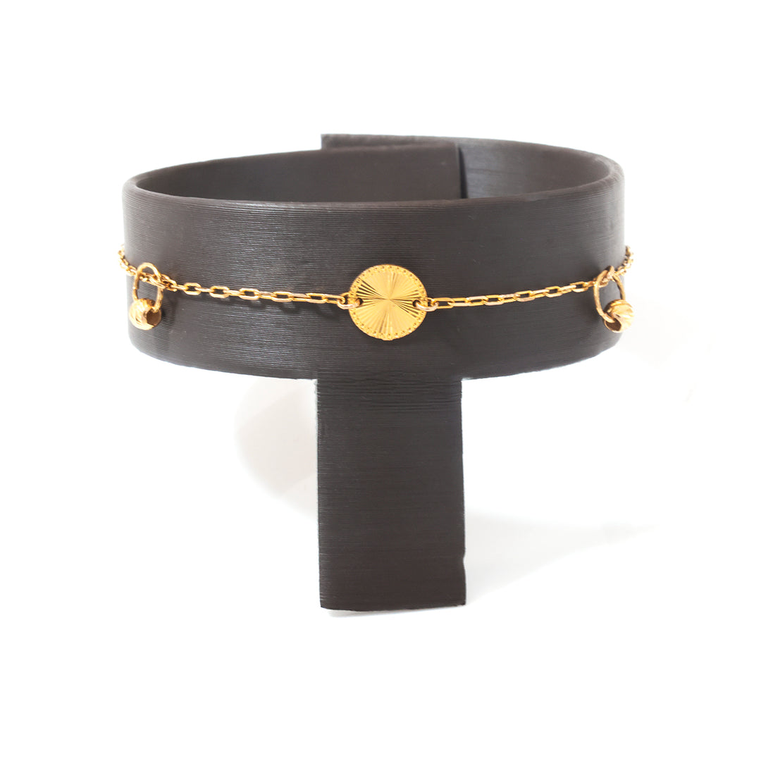 A gold chain bracelet with a round textured disc charm and small dangling beads, displayed on a black T-shaped jewelry stand against a white background.