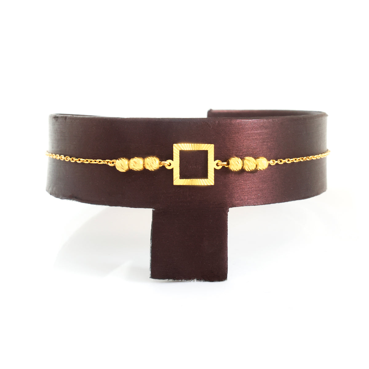 A wide dark brown leather cuff bracelet with a gold chain, gold square buckle, and small gold beads, displayed on a black stand against a white background.
