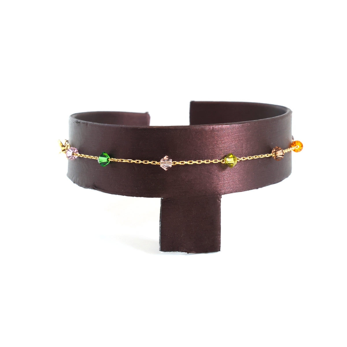 A dark brown leather cuff bracelet with a delicate gold chain studded with small colorful gemstones, displayed on a white background.
