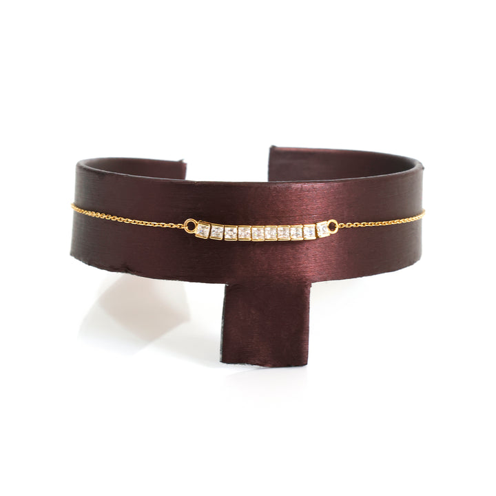 A wide dark brown leather cuff bracelet with a delicate gold chain running horizontally across the front, featuring a small gold bar set with clear rectangular gemstones in the center.