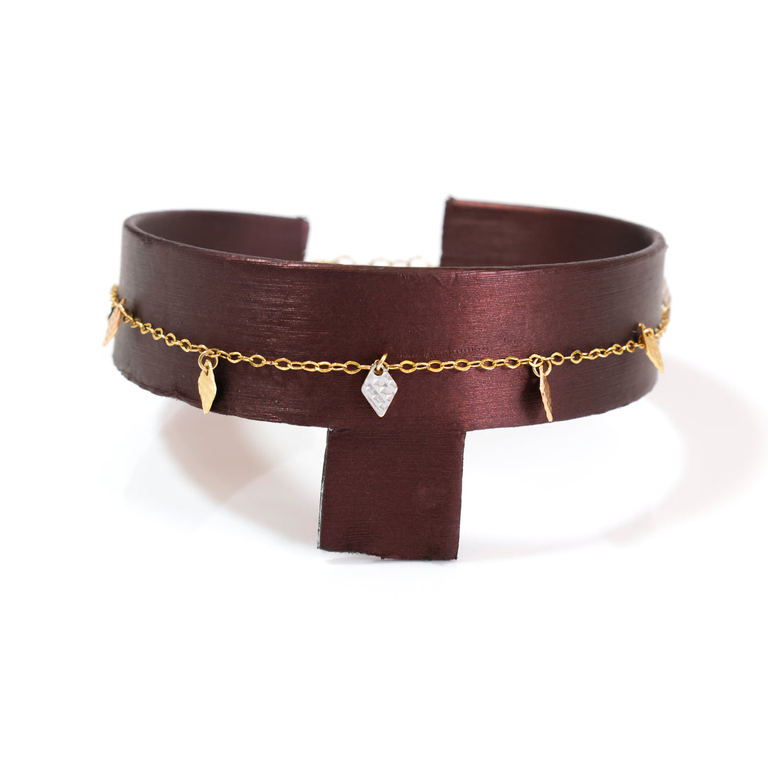 A dark brown leather cuff bracelet with a delicate gold chain draped across the front, adorned with small gold leaf charms and a clear faceted crystal pendant, displayed on a white background.