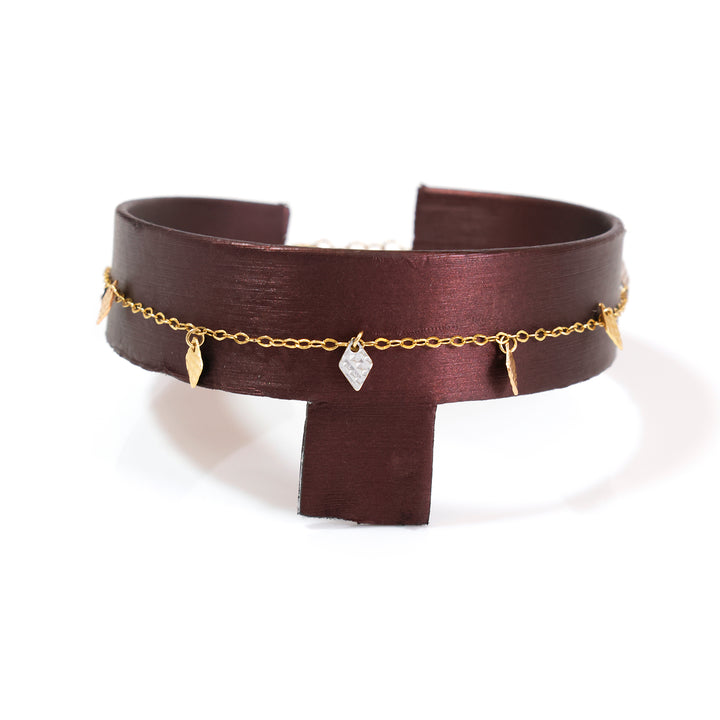 A dark brown leather cuff bracelet with a delicate gold chain draped across the front, adorned with small gold leaf charms and a clear faceted crystal pendant, displayed on a white background.