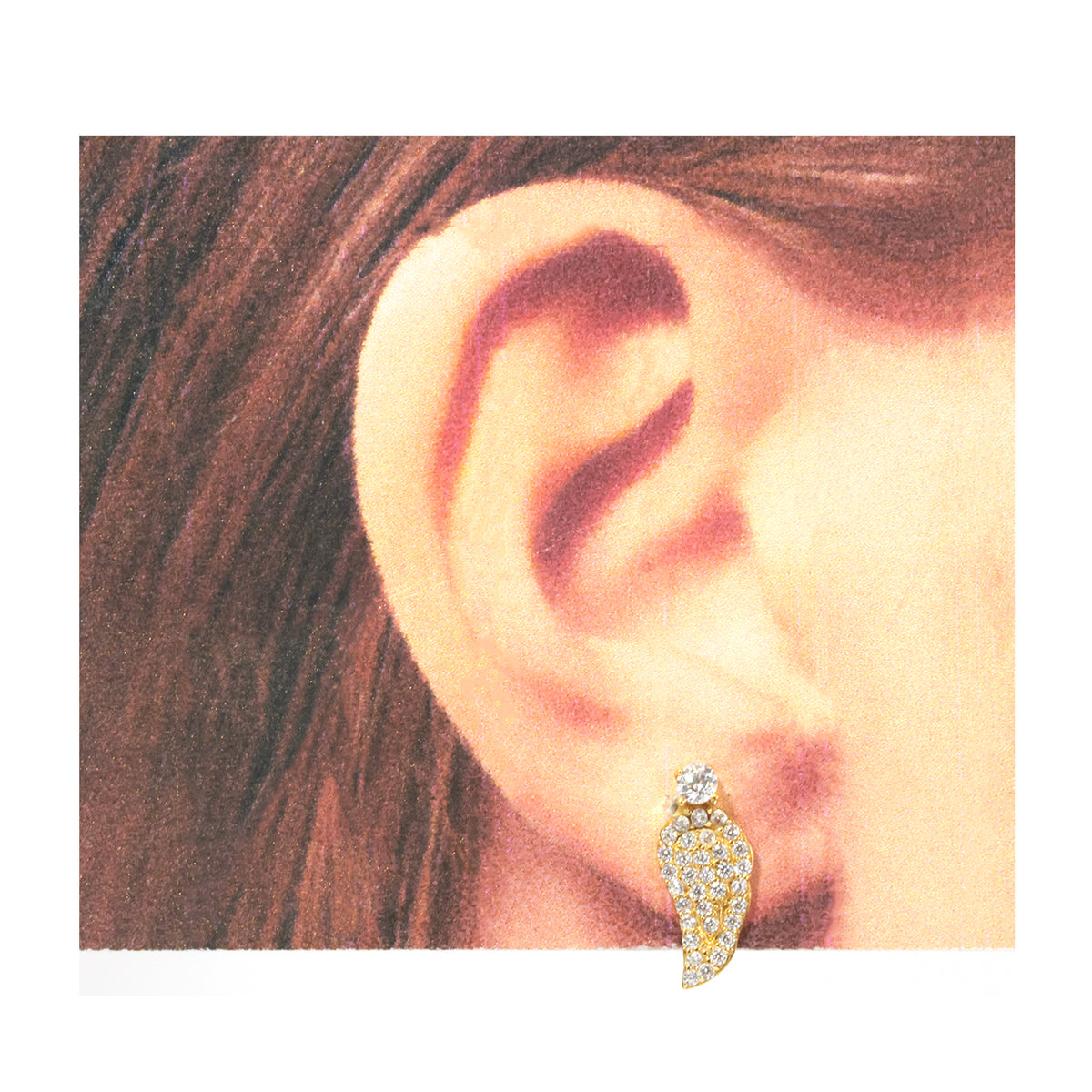 A close-up of a person's ear wearing a small wing-shaped stud earring. The earring is gold-toned with clear pavé stones, and the surrounding background is mostly white with a warm-toned inset photo.
