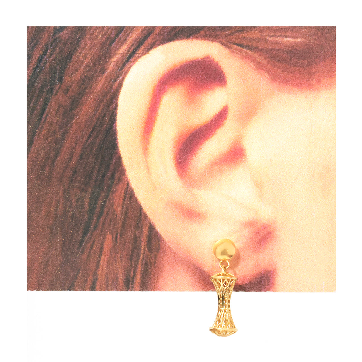 A gold dangle earring with a textured filigree drop and a round stud, shown worn on a person's ear against a white background.