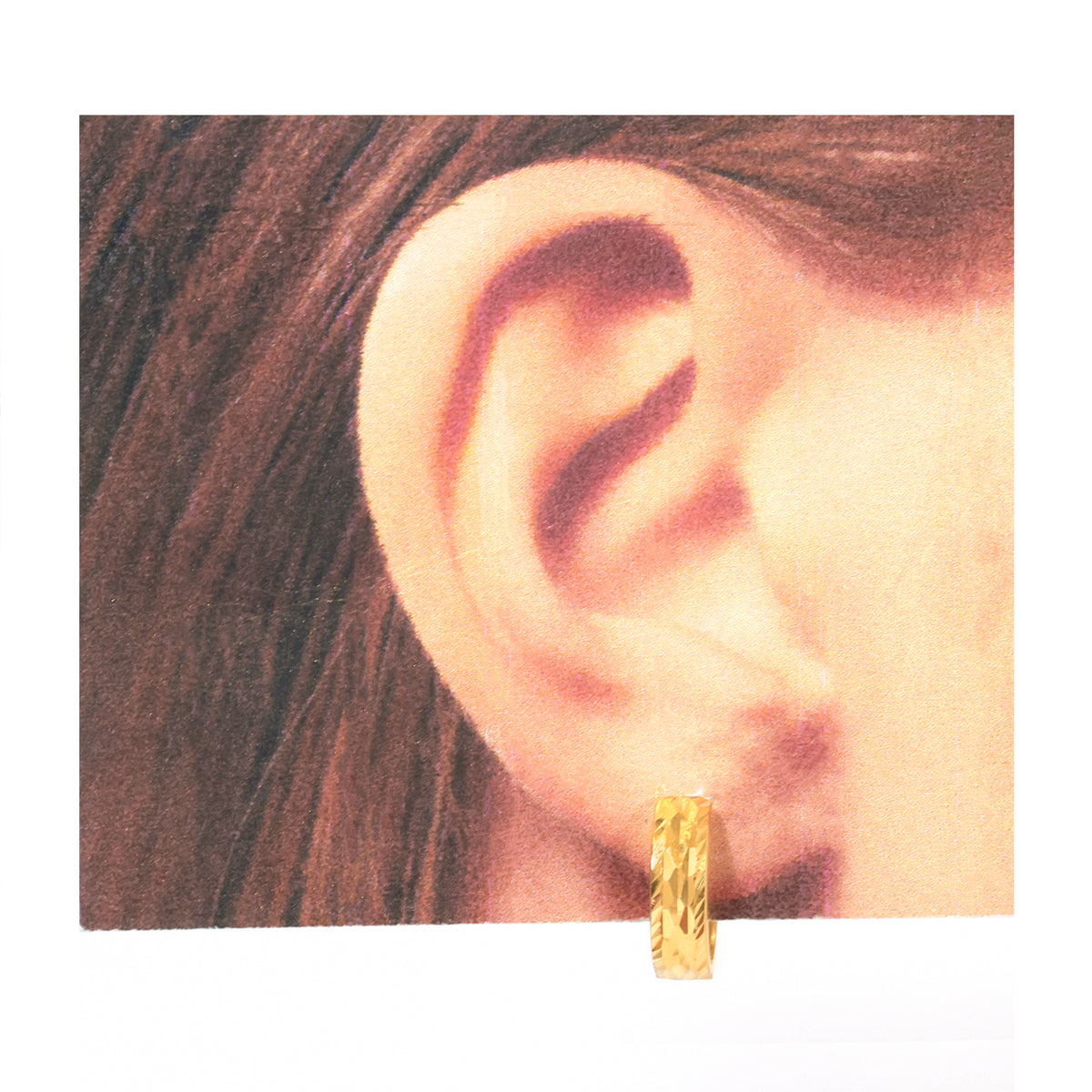 A close-up of a person's ear wearing a small textured gold hoop earring, shown against a white background with a printed card behind the ear.