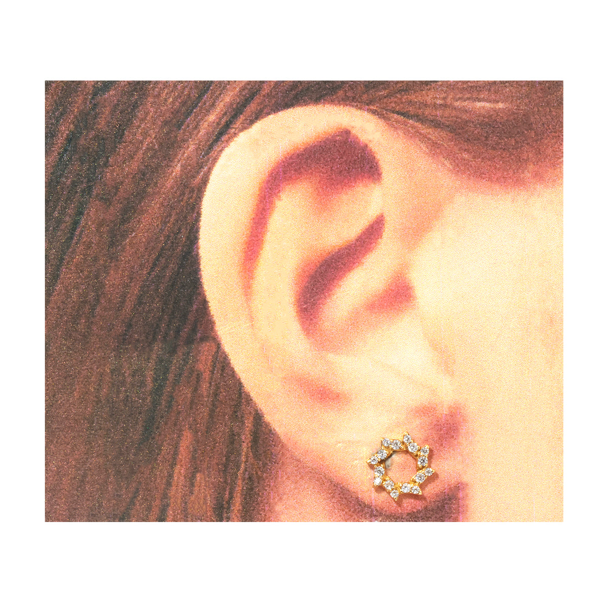 A close-up of a person's ear wearing a small gold-toned stud earring with multiple clear stones, shown against a white border.