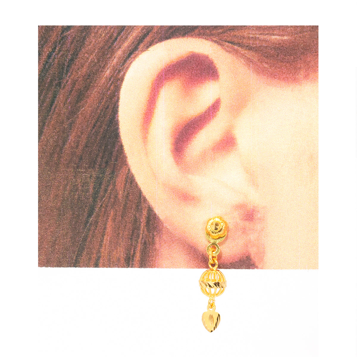 A gold dangle hoop earring with a textured top ball, a small chain, a faceted bead, and a dangling heart charm, shown worn on a model's ear against a white background.