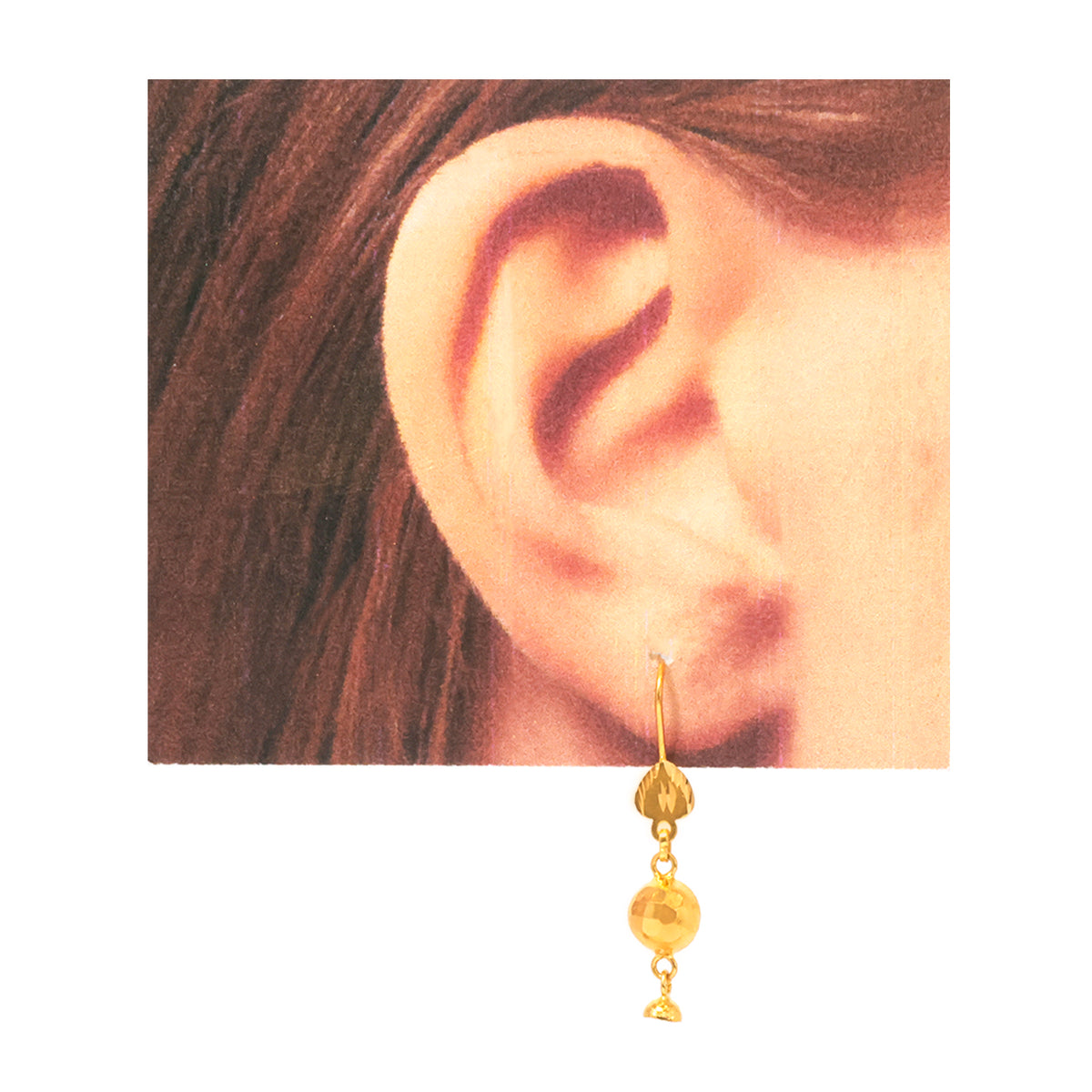 A close-up of a person's ear wearing a small gold dangle earring with two hanging gold beads and a tiny bell-shaped charm, shown against a plain white background.