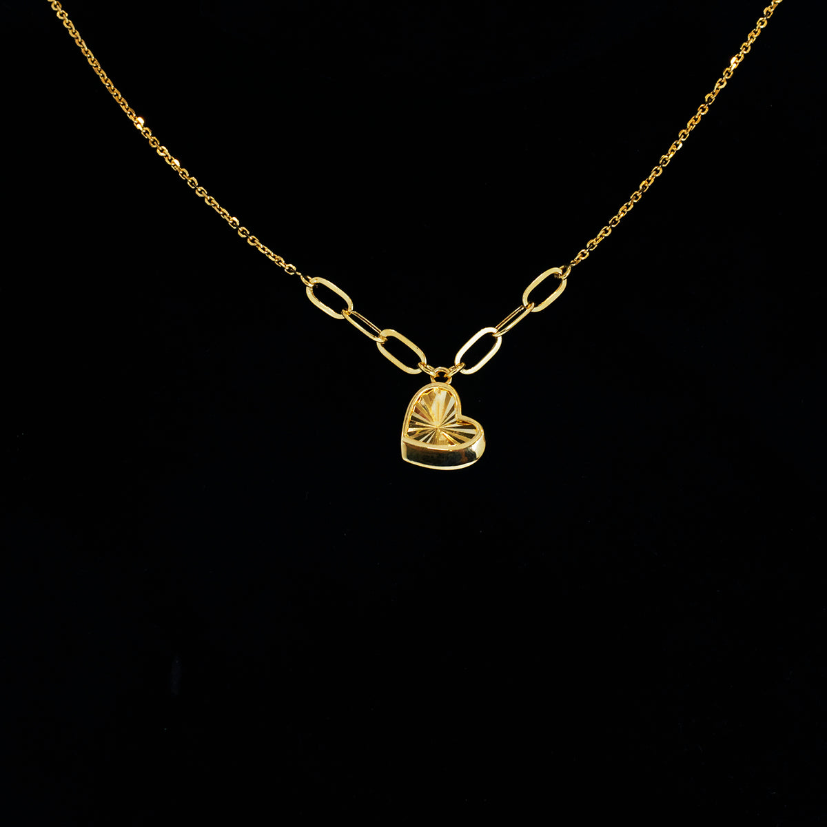 A gold chain necklace featuring a paperclip-style connector leading to a small heart-shaped pendant with a sunburst or fan-like textured design, displayed against a solid black background.