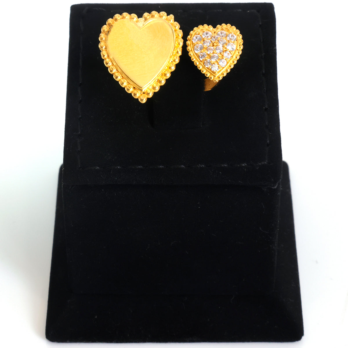 A pair of heart-shaped gold rings displayed on a black velvet jewelry stand against a white background. One ring features a smooth, polished gold heart with a beaded border, while the other features a gold heart paved with small clear stones.