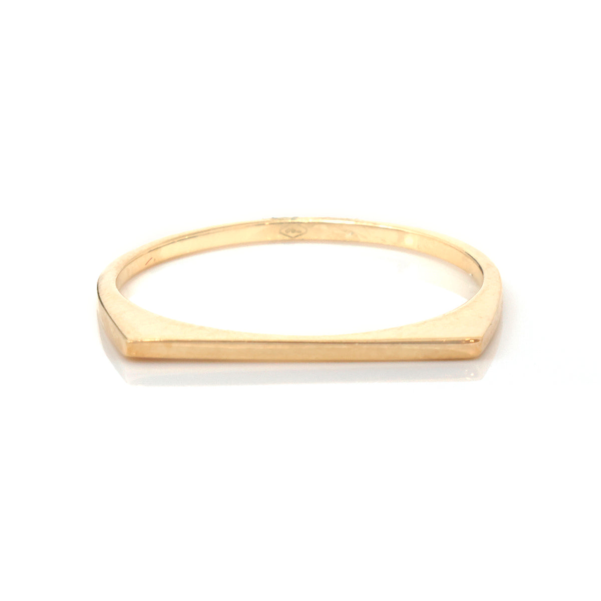 A minimalist gold ring with a flat, geometric, slightly tapered band, photographed on a clean white background with a soft reflection.