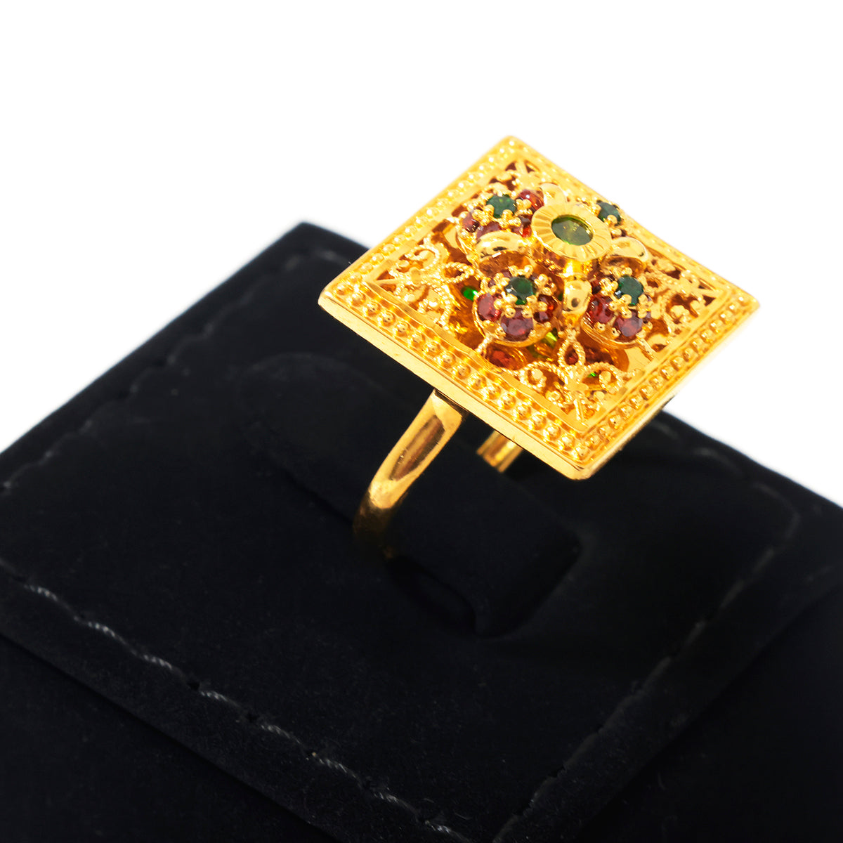 A gold square ring with intricate filigree detailing and small red, green, and blue gemstones, displayed on a black velvet ring box against a white background.