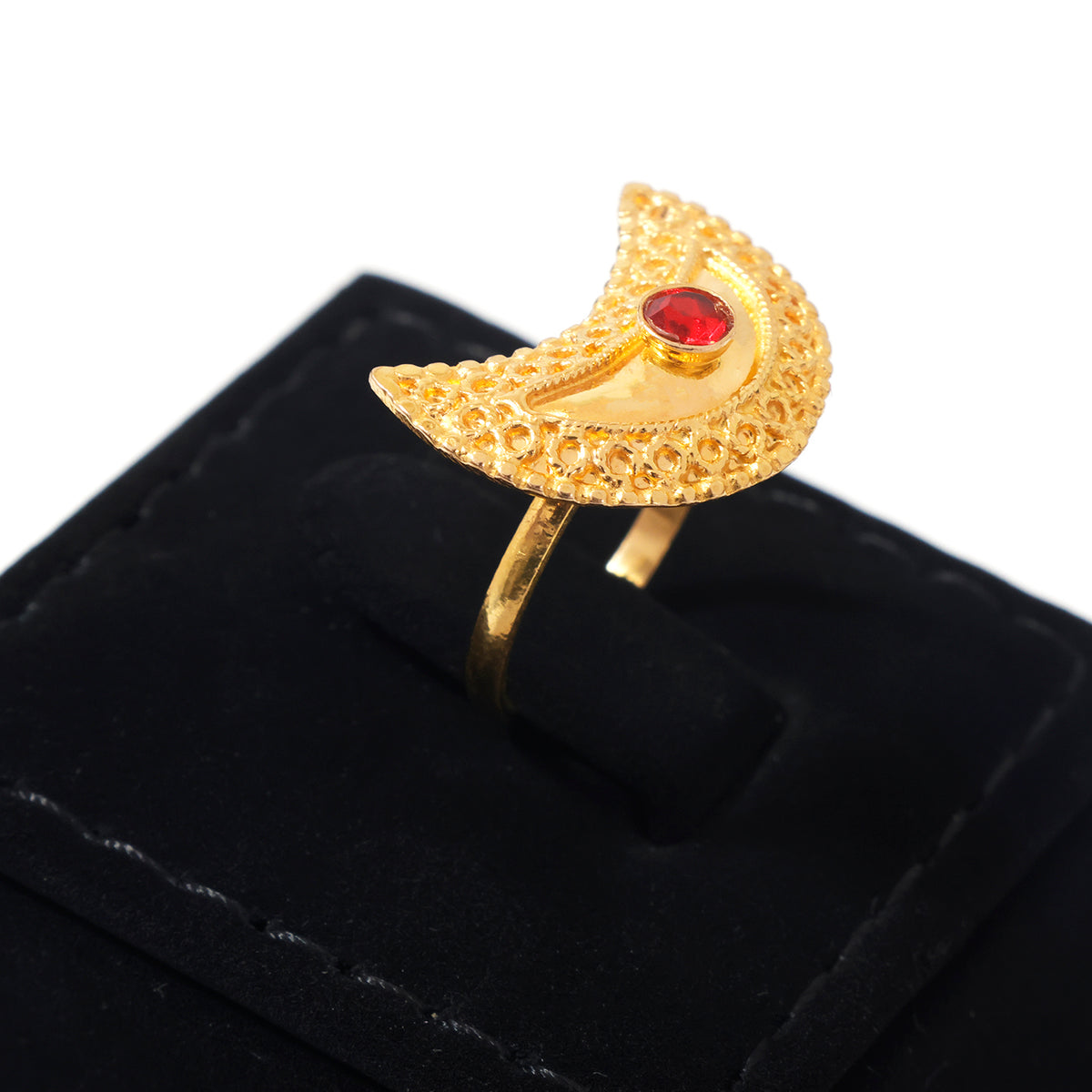 A gold crescent moon ring with a textured filigree design and a central red gemstone, displayed on a black velvet ring box against a white background.