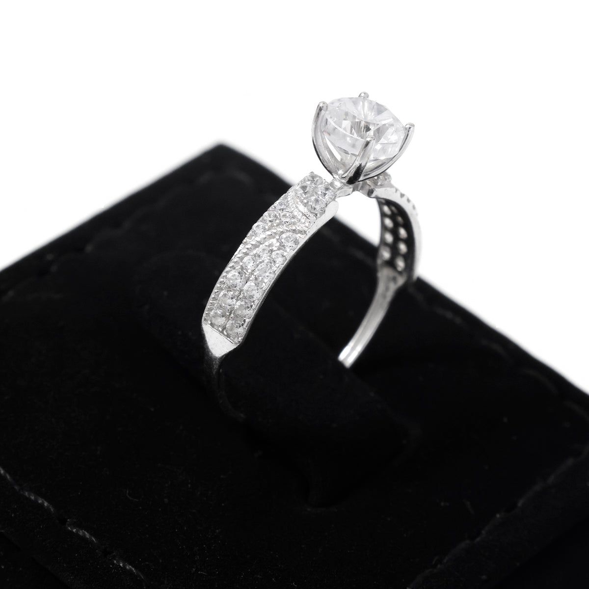 A silver-toned diamond engagement ring with a round-cut center stone and pavé-set side stones, displayed on a black velvet ring box against a white background.