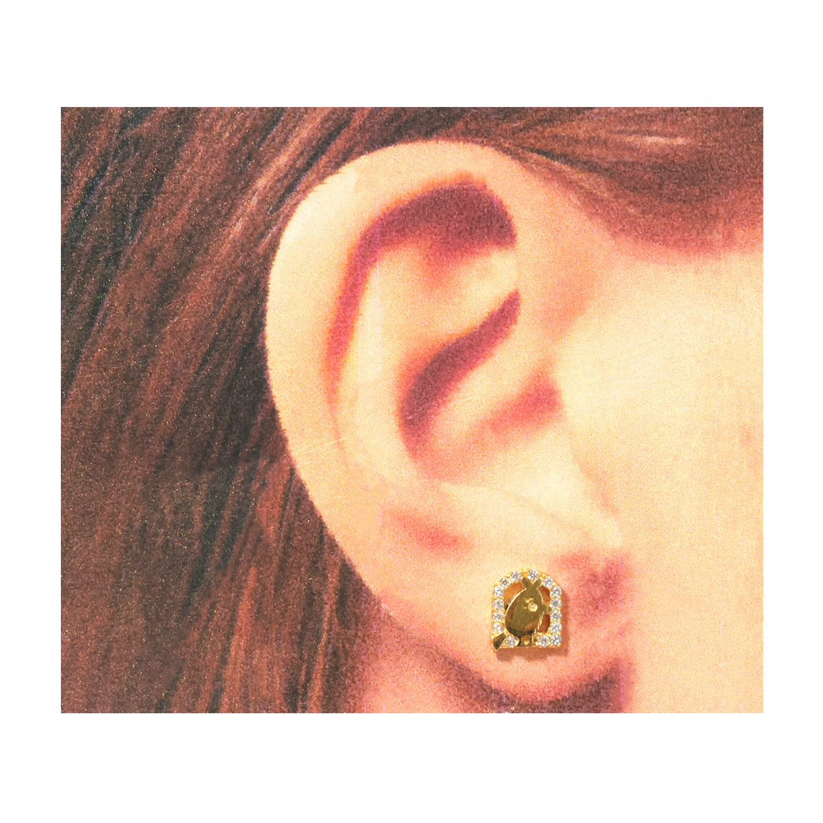 A close-up of a person's ear wearing a small gold stud earring with a central oval stone and a halo of clear stones.