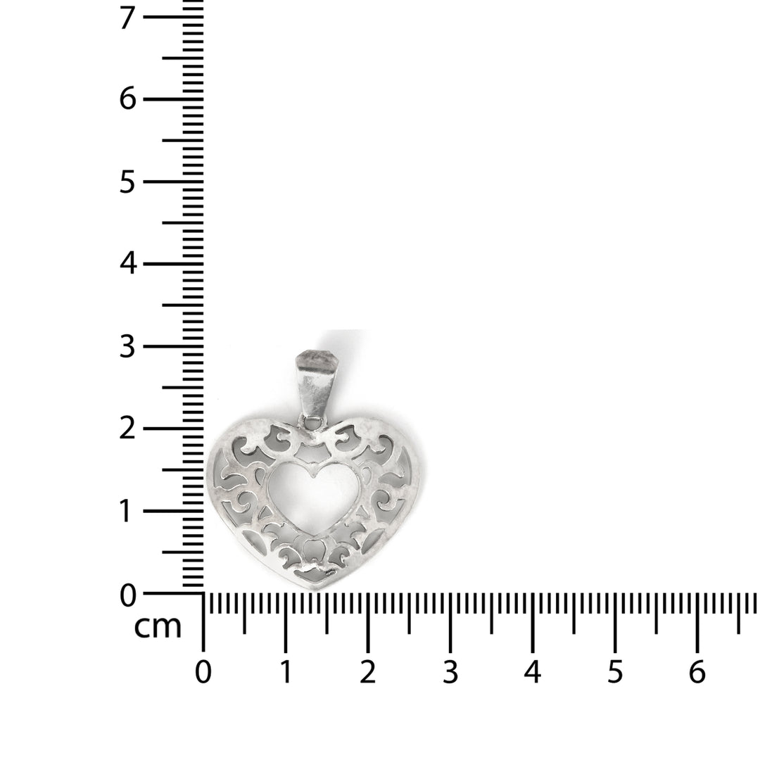 Silver heart-shaped pendant with intricate designs on a white background with a ruler for scale.