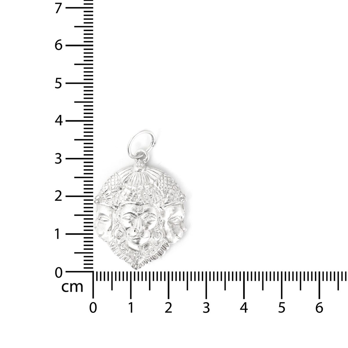 Silver pendant with face design next to a ruler for scale on a white background